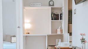 Une pièce compacte et bien éclairée, comprenant un lit, une table à manger avec des chaises, une petite bibliothèque, une horloge et un téléviseur fixé au mur.
