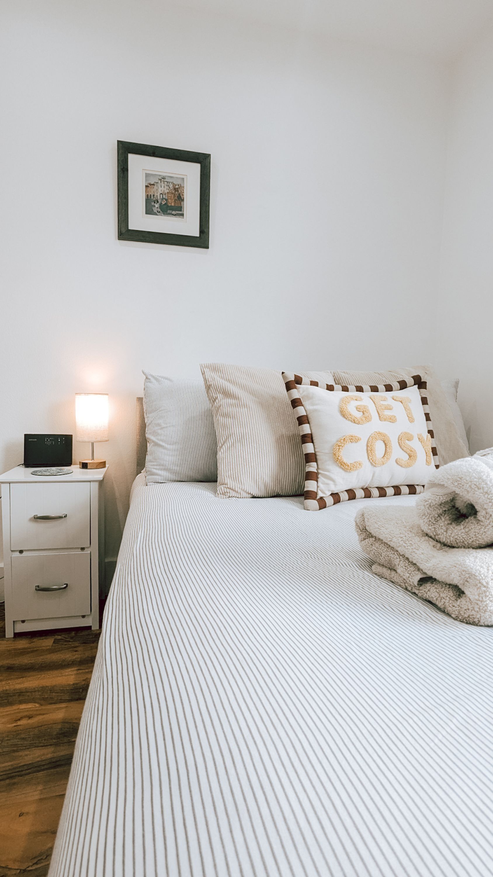 Een keurig opgemaakt slaapkamer met een bed, kussens, een nachtkastje met een lamp en een ingelijste foto aan de muur.