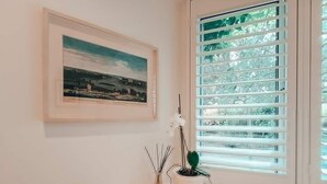 Ein Zimmer mit einem gerahmten Landschaftsbild, einem Fenster mit Jalousien und einem kleinen Tisch mit Blumentopf und einem Strauß Zweige.