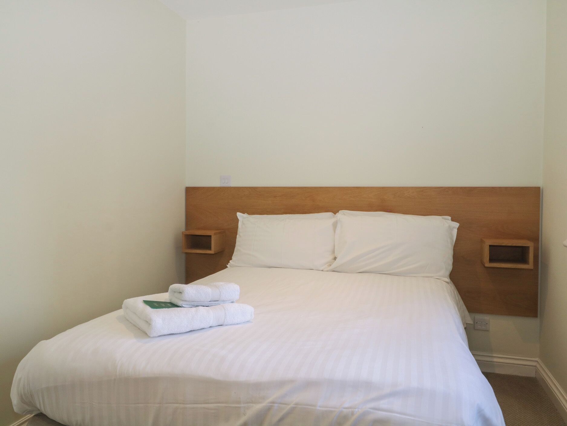 A neatly made bed with white linens and pillows, two bedside tables with wooden headboards, and a small green sign on the bed.