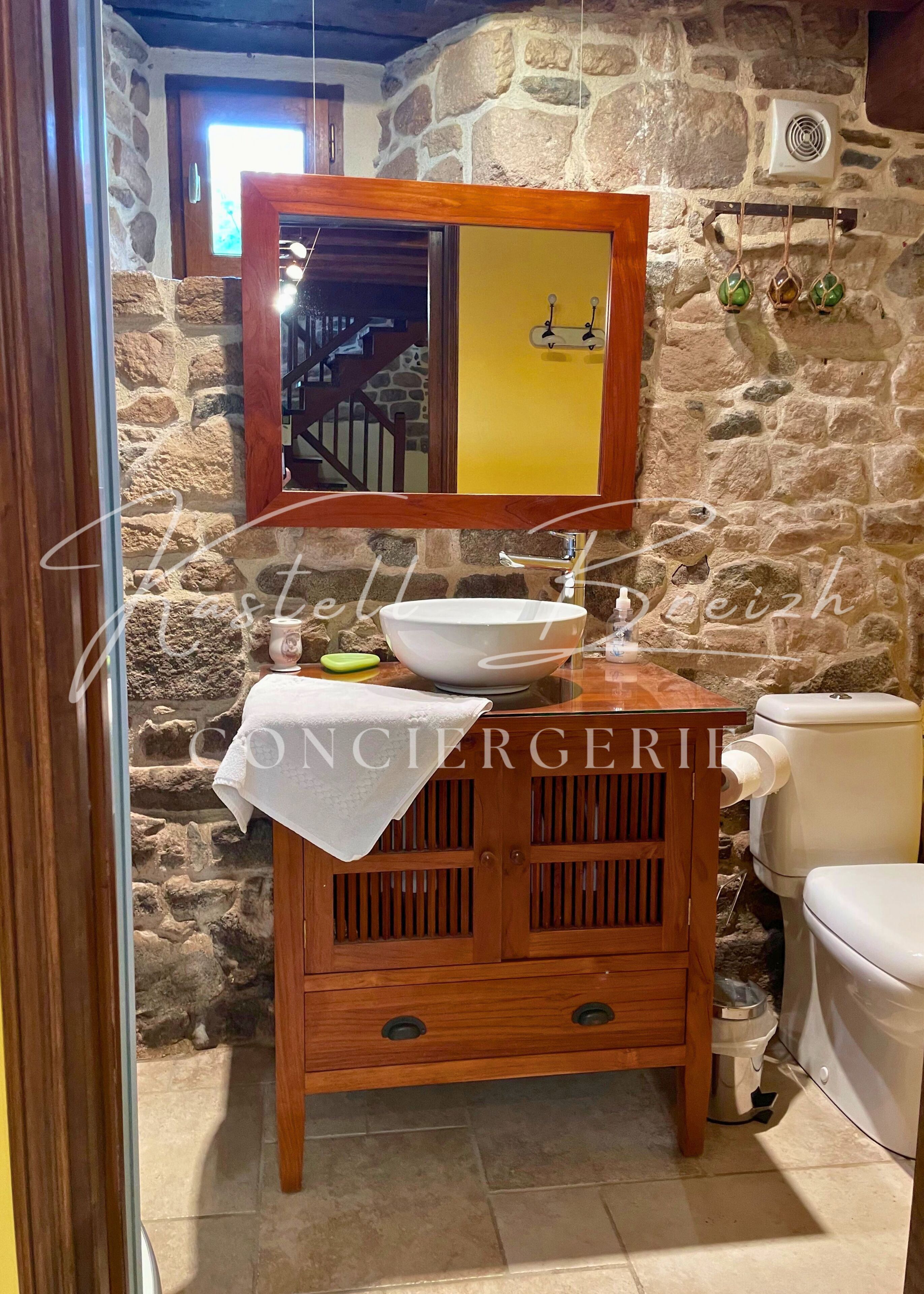 Un baño con pared de piedra, tocador de madera con lavabo, espejo y retrete.