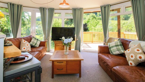 A cozy living room with leather sofas, a wooden coffee table, and a television. Large windows offer a view of a wooden deck and greenery.