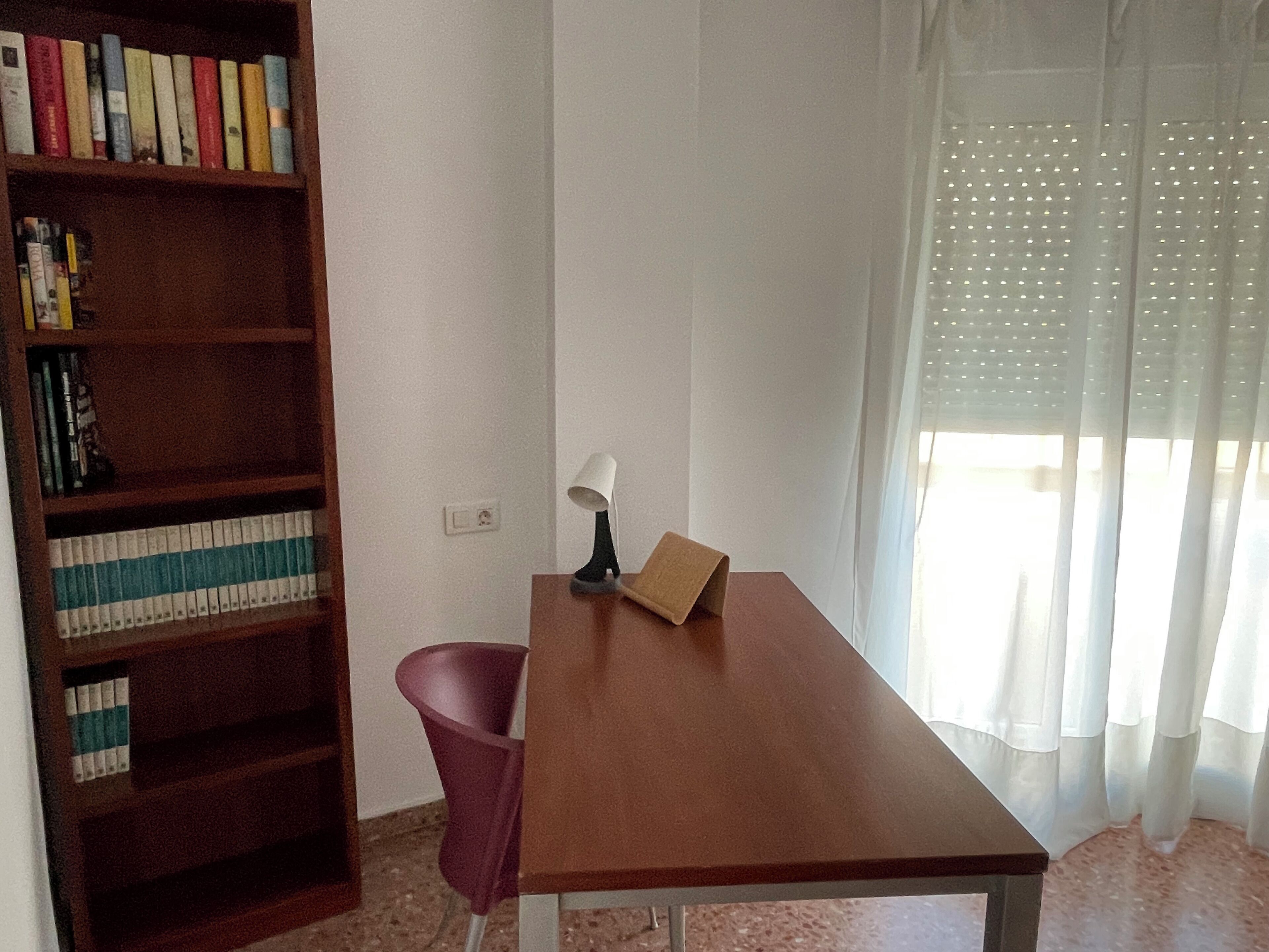A room with a wooden desk, a chair, a bookshelf, and a window with sheer curtains.