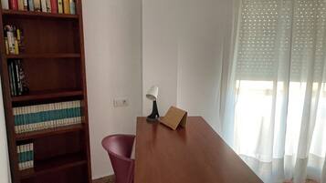 A room with a wooden desk, a chair, a bookshelf, and a window with sheer curtains.