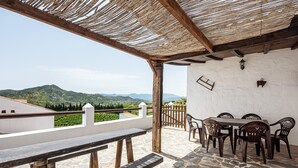 Une terrasse extérieure avec un sol en pierre, des bancs en bois, une table avec des chaises, un toit de chaume et une vue sur des collines verdoyantes.