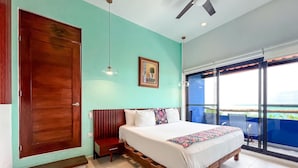 Une chambre avec un grand lit, une armoire en bois, un ventilateur de plafond, un tableau encadré au mur et une vue sur le balcon extérieur.