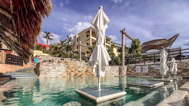 Une piscine aux eaux cristallines, agrémentée de marches en pierre, de parasols blancs et d’un mur de pierre sur lequel s’élève un escalier.