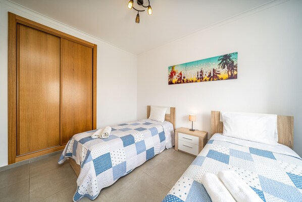 A bedroom with two beds, a wooden wardrobe, a nightstand, a lamp, and a wall painting.