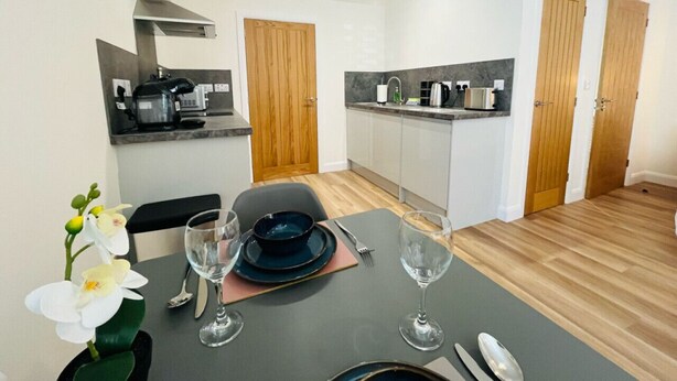 A modern kitchen with white cabinets, a built-in oven, and a microwave. There is a dining area with a table set for two, including plates, glasses, and cutlery. - Viewforth Studio No.4 (Fort William)