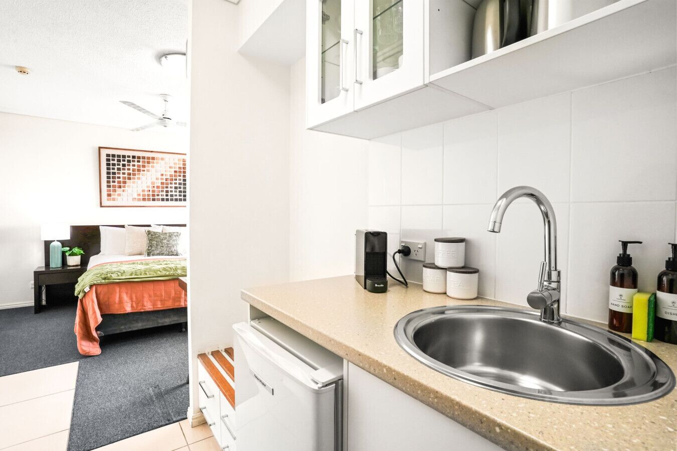 A modern hotel room with a bed, bedside table, and a kitchenette area including a sink and cabinets.
