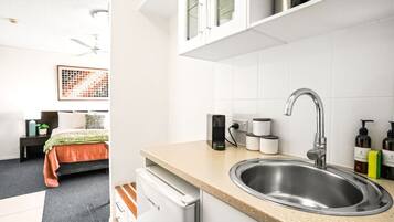 A modern hotel room with a bed, bedside table, and a kitchenette area including a sink and cabinets.