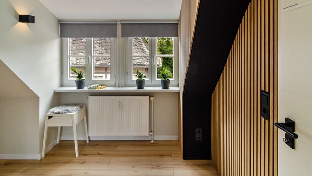Une pièce avec un radiateur, une chaise blanche, une fenêtre ornée de plantes et un mur recouvert de panneaux de bois.