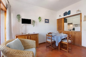 A room with a wicker chair, a dining table with a striped tablecloth, a television on the wall, and a kitchen area with cabinets and a mirror.