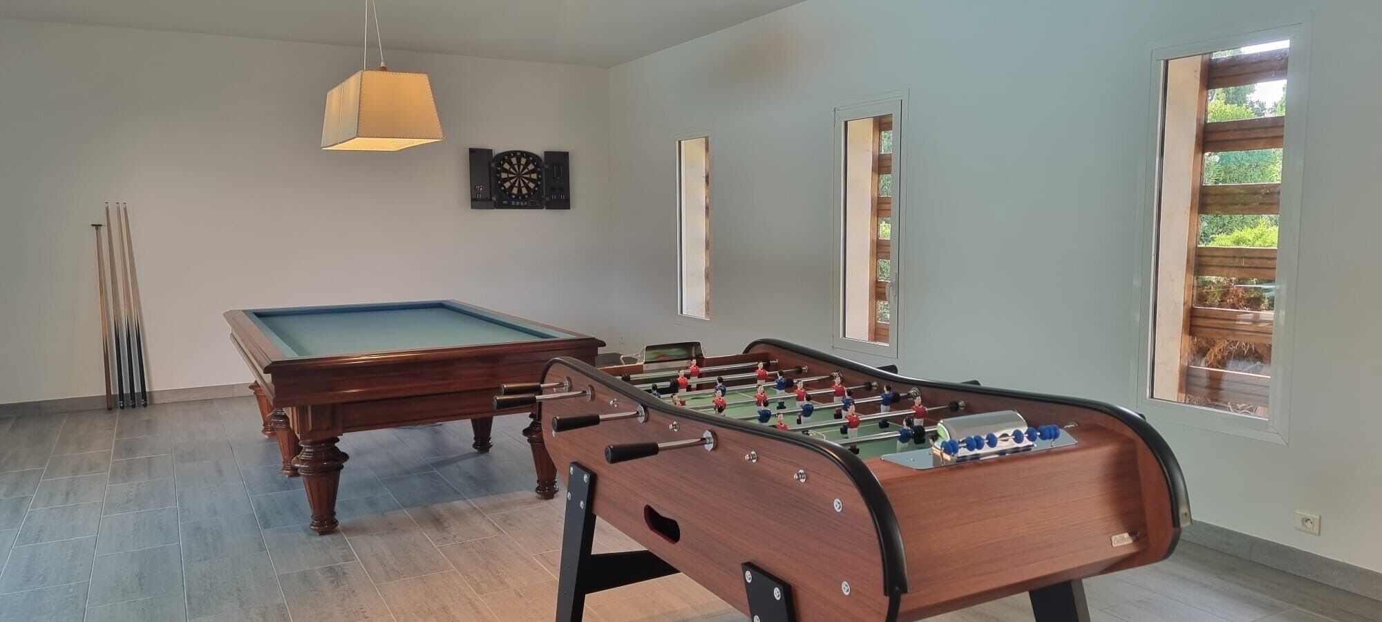 Ett biljardbord, ett fotbollsspel och en uppsättning pilar i ett rum med stora fönster och en hängande lampa.