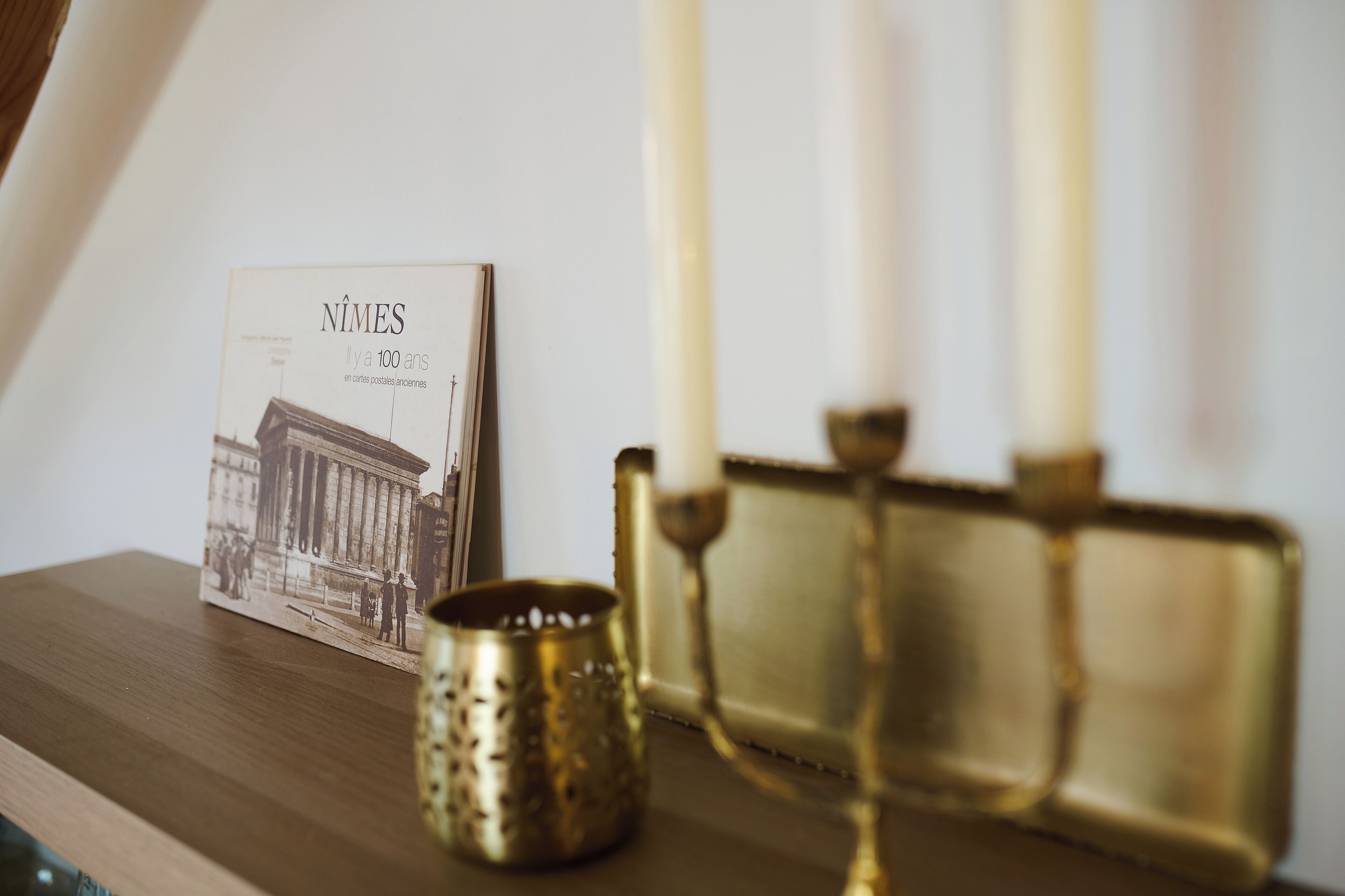 A framed book titled "NIMES" on a wooden surface, a brass candle holder with a candle, and a brass candelabra with candles.