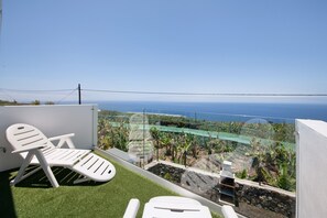 A balcony with a white chair, a small table, and a view of the ocean and garden.