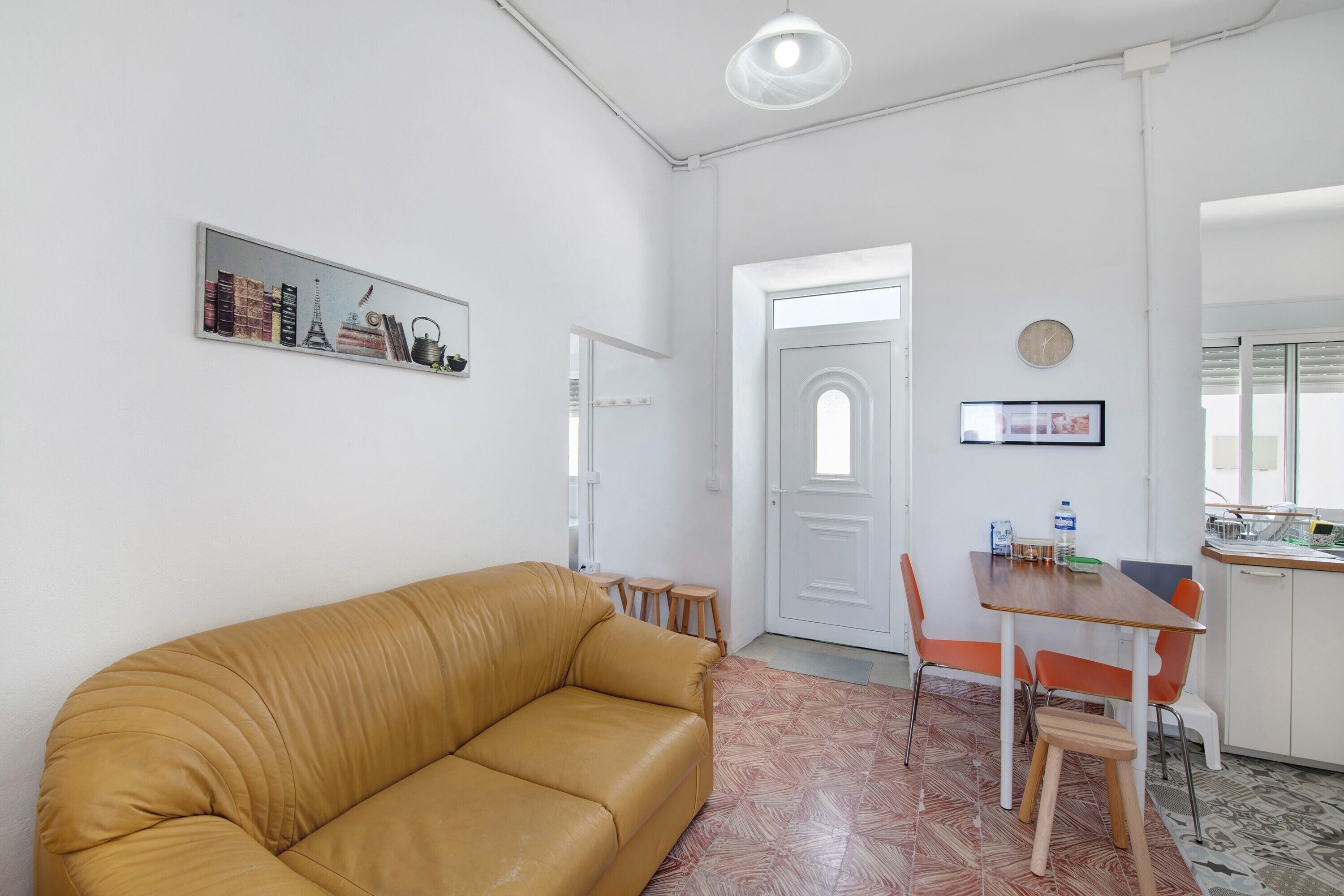 A living room with a tan sofa, a dining table with chairs, a kitchen area, and a white door.
