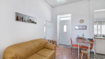 A living room with a tan sofa, a dining table with chairs, a kitchen area, and a white door.