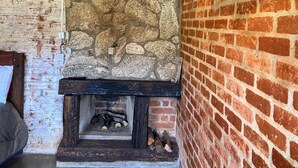 Une cheminée en pierre avec un manteau en briques, des poutres en bois au plafond et un lit avec une tête de lit située à gauche.