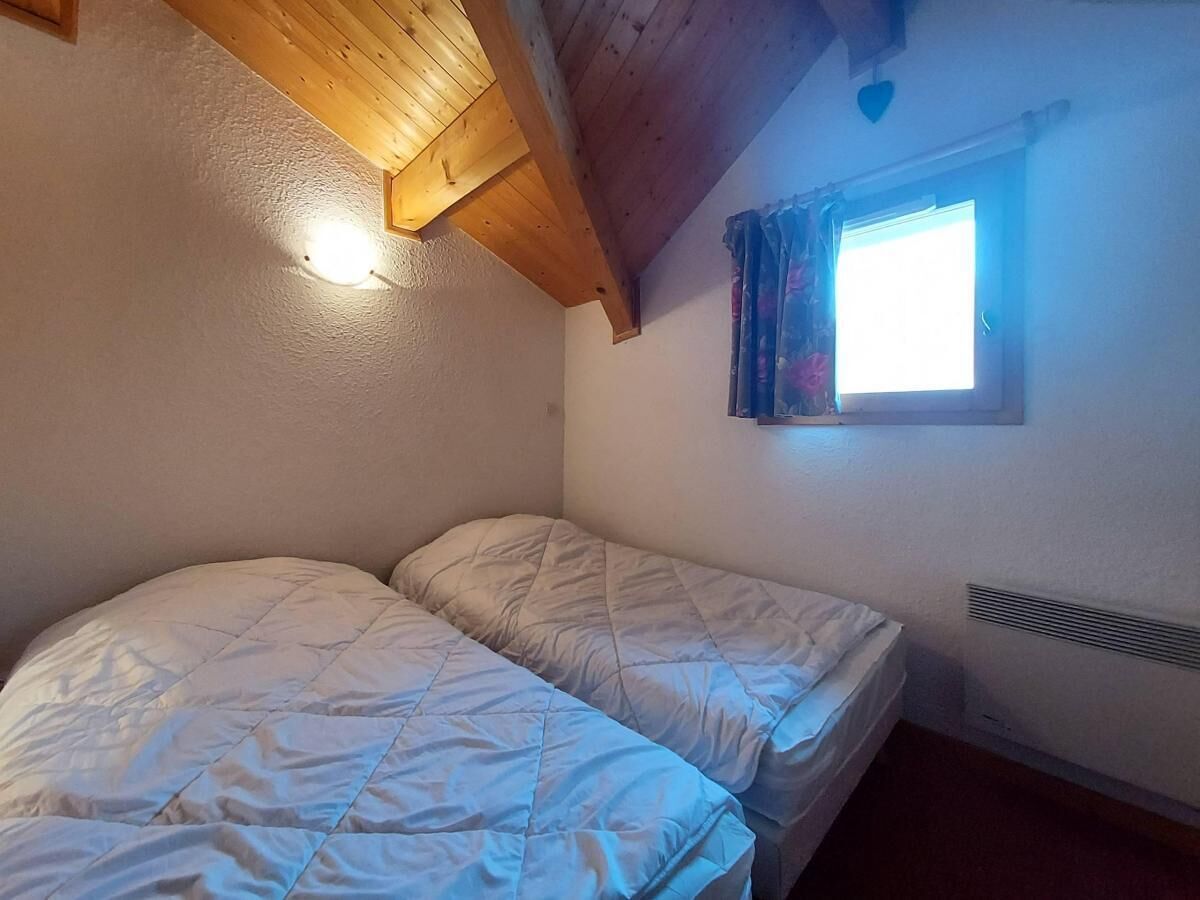A small, cozy room with two single beds, a sloped ceiling, a window with curtains, and a wall-mounted heater.