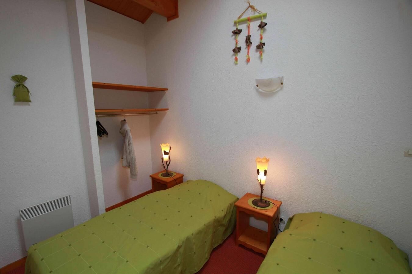 A small, cozy room with two single beds, a wooden shelf, a wall-mounted hanging, and a radiator.