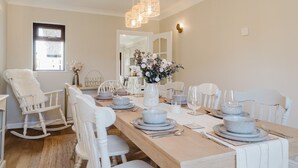A dining room with a long table set for six, white chairs, a white rocking chair, a vase with flowers, and hanging wicker lamps.