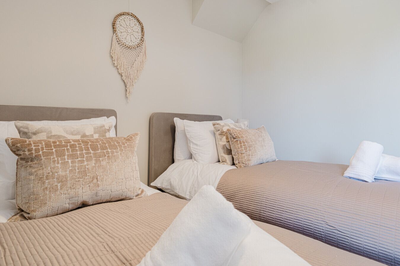 Une chambre d’hôtel avec deux lits, chacun recouvert d’une couvre-lit beige et agrémenté d’oreillers, une décoration murale en macramé et un lit parfaitement fait avec une serviette blanche.