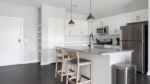 A modern kitchen with white cabinets, a stainless steel refrigerator, a microwave, and a sink with a faucet. There are two bar stools at the island. - Landing | Beautiful 2BD, Gym, Pool (Mableton)