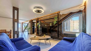 Un salon moderne avec des canapés bleus, une table basse ronde en bois et un téléviseur fixé sur un mur foncé. Des plantes sont disposées sur des étagères, et une grande fenêtre offre une vue sur les bâtiments environnants.