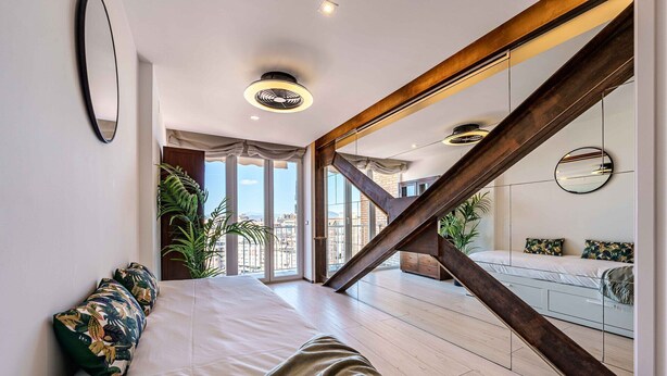 Une chambre moderne avec un grand lit, un escalier en bois et une vue sur la ville grâce à de grandes fenêtres.