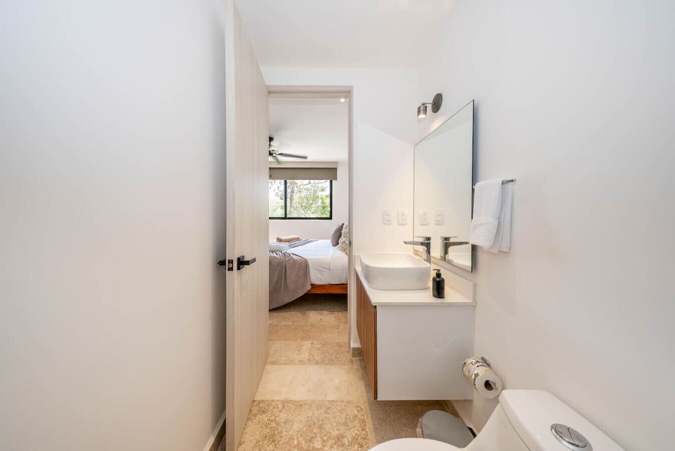 A modern bathroom with a sink, mirror, and toilet. A bed with a window is visible in the background.