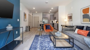 A modern living room with a sofa, coffee table, and a blue rug. There is a kitchen in the background with cabinets and appliances. - Landing | Chic 1BD, Yoga Studio, Gym (Santa Rosa Beach)