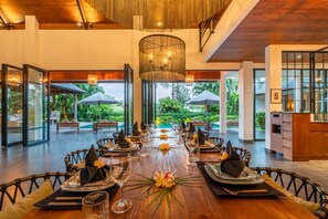 A dining area with a long wooden table set for a meal, surrounded by chairs with cushions. - Infinity pool - 4 King Beds - Ricefield View (Canggu)