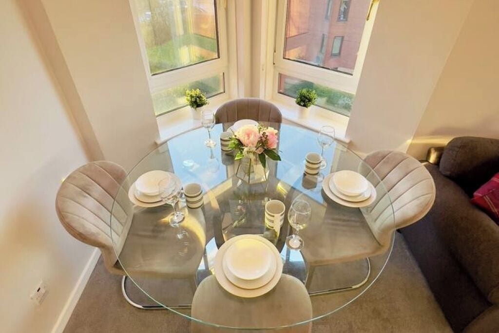 A round glass table set for four with a vase of flowers, positioned in a room with a view of a building through large windows.