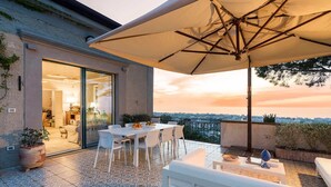 Un patio al aire libre con una mesa y sillas, un paraguas grande y vistas a la ciudad al atardecer.