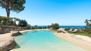 Una piscina cristalina con borde arenoso, rodeada de rocas y plantas, con vista al océano y un cielo despejado.
