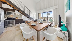 A modern kitchen with a microwave, stove, and cabinets. There is a dining area with a wooden table and white chairs. A staircase leads to a balcony with a view.