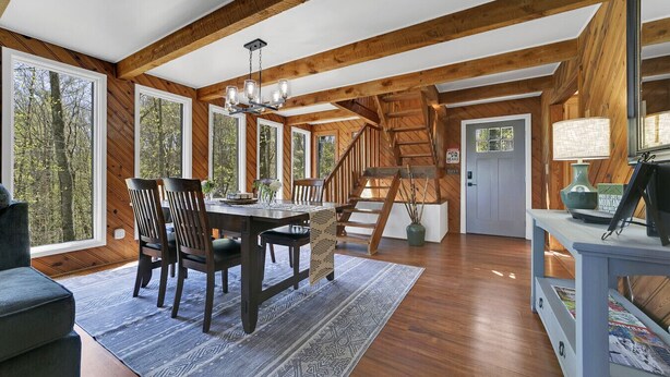 A spacious room with a wooden staircase, a dining area with a table and chairs, and large windows offering a view of the outdoors.