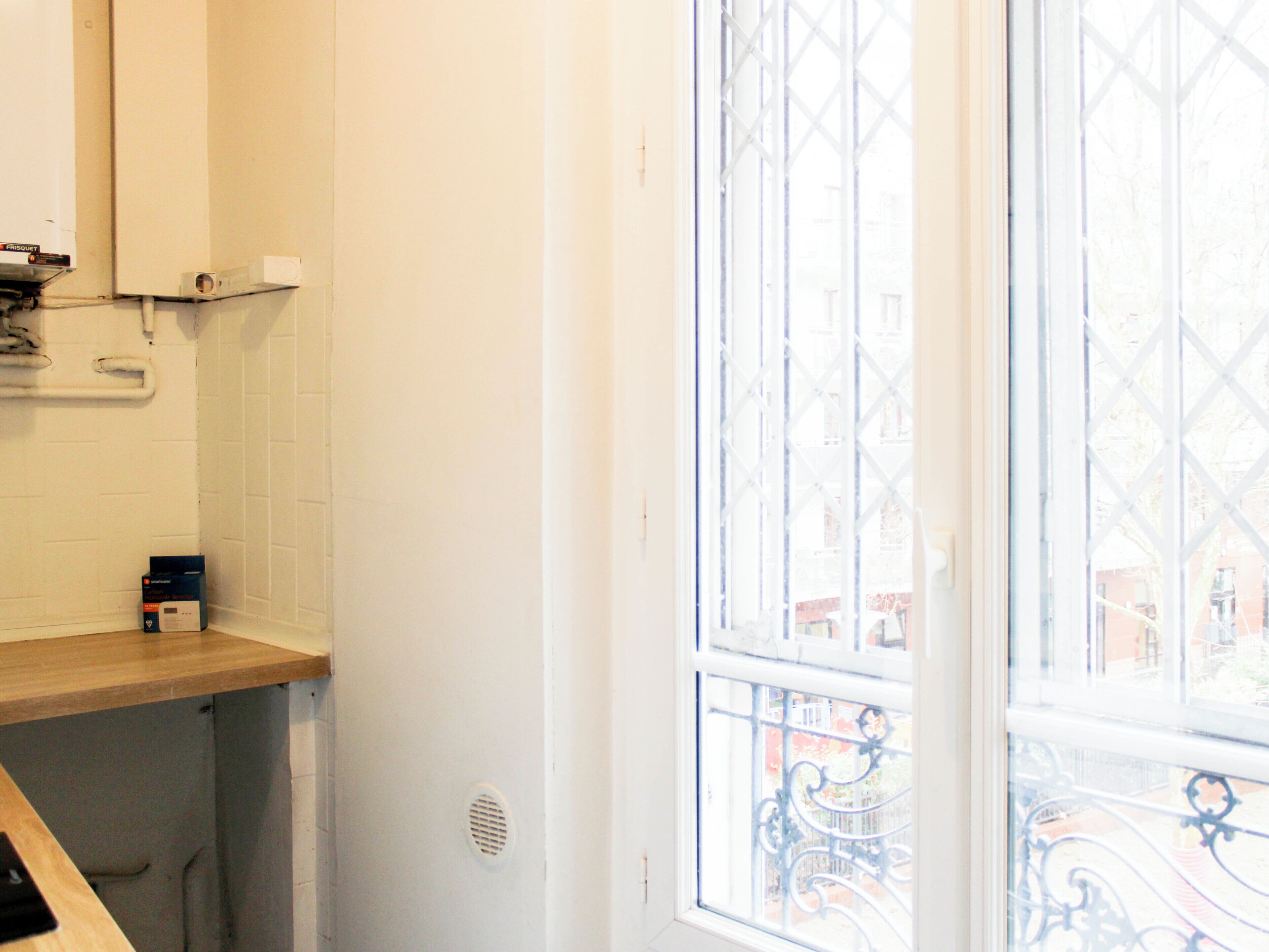 Une salle de bain avec un plan de toilette en bois, un mur blanc, une fenêtre avec une grille en métal et une bouche d’aération visible.
