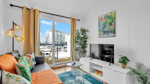 A modern living room with a sofa, TV, and a view of a cityscape through large windows. - On the Water 6 with Free Parking | By My Getaways (Brighton)