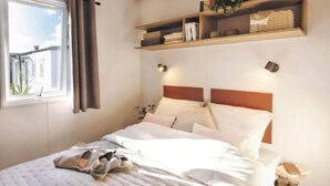 A neatly made bed with pillows and a handbag, a bedside table with a lamp, and a window with curtains. - ZEcamping 3* pet friendly mobil-home TV LV clim 32m2 6 pers (Bauduen)