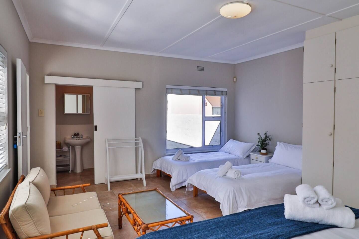 A hotel room with two beds, a sitting area with a chair and table, a bathroom with a sink and mirror, and a large window.