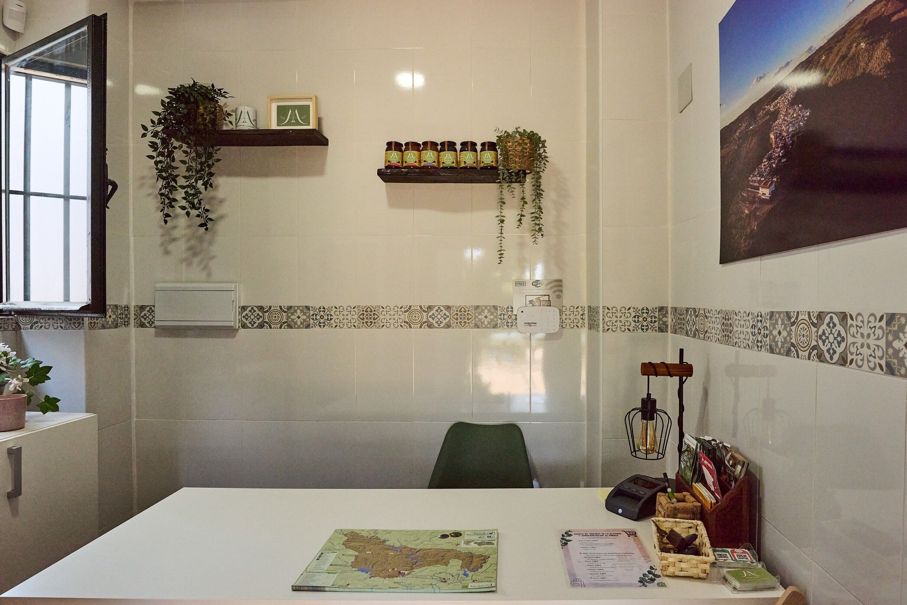 A room with a map, a desk, a chair, a shelf with bottles, a hanging plant, and a wall-mounted light fixture.