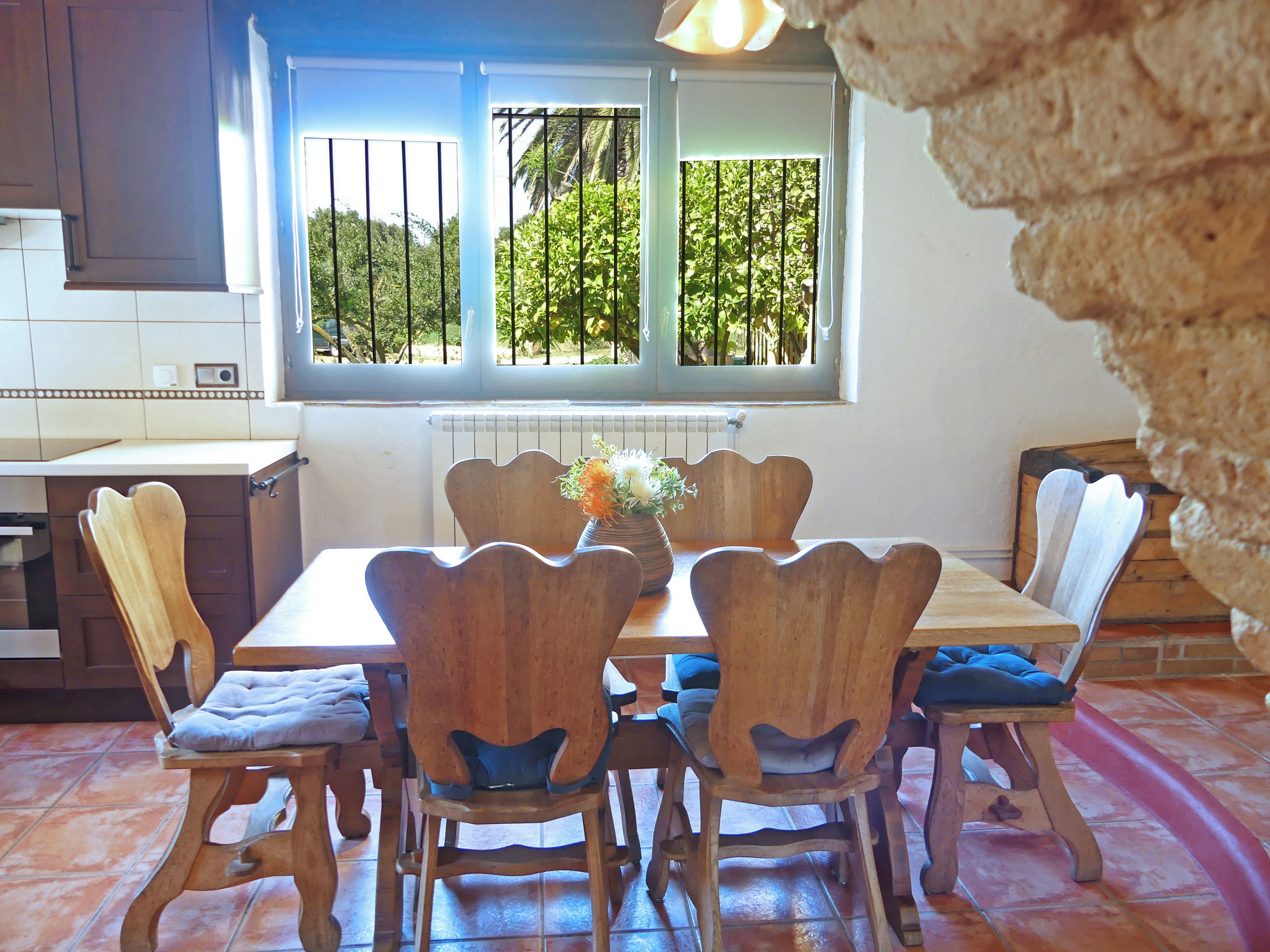 Un comedor con una mesa y sillas de madera, una ventana con vista a follaje, y una cocina al fondo.