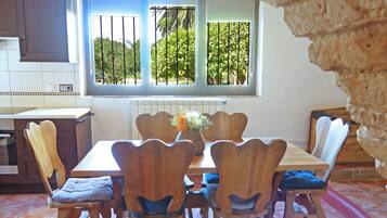Un comedor con una mesa y sillas de madera, una ventana con vista a follaje, y una cocina al fondo.