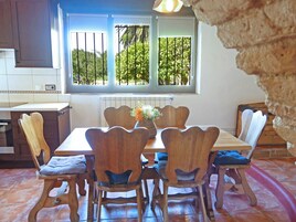 Un comedor con una mesa y sillas de madera, una ventana con vista a follaje, y una cocina al fondo.