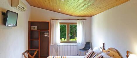 A bedroom with a wooden ceiling, a bed with striped bedding, a window with curtains, a radiator, a TV on the wall, and a small table with a lamp and flowers.