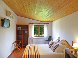 Un dormitorio con techo de madera, una cama con ropa de cama a rayas, una ventana con cortinas, un radiador, un televisor en la pared y una mesita con una lámpara y flores.
