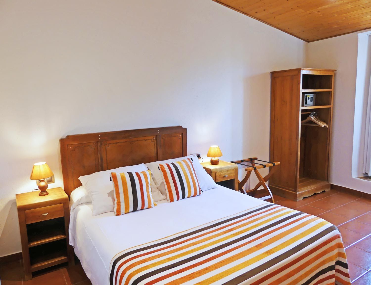 Un lit bien fait, avec une literie à rayures, une tête de lit en bois, une table de chevet avec une lampe et une armoire en bois dotée d’une étagère pour suspendre des vêtements.