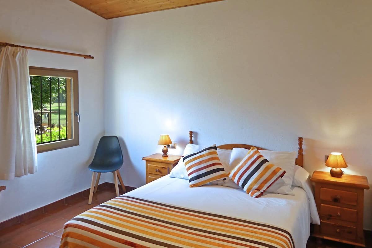 Une chambre à coucher avec un lit, deux tables de chevet avec des lampes, une chaise, une fenêtre avec des rideaux et une vue sur la verdure à l’extérieur.